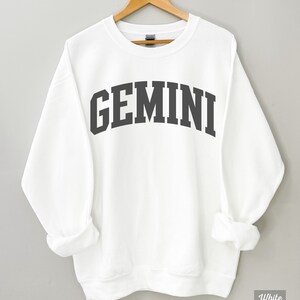 以下が含まれることがあります： 白地に黒で「GEMINI」とプリントされたスウェットシャツ。大学風のフォントを使用しています。