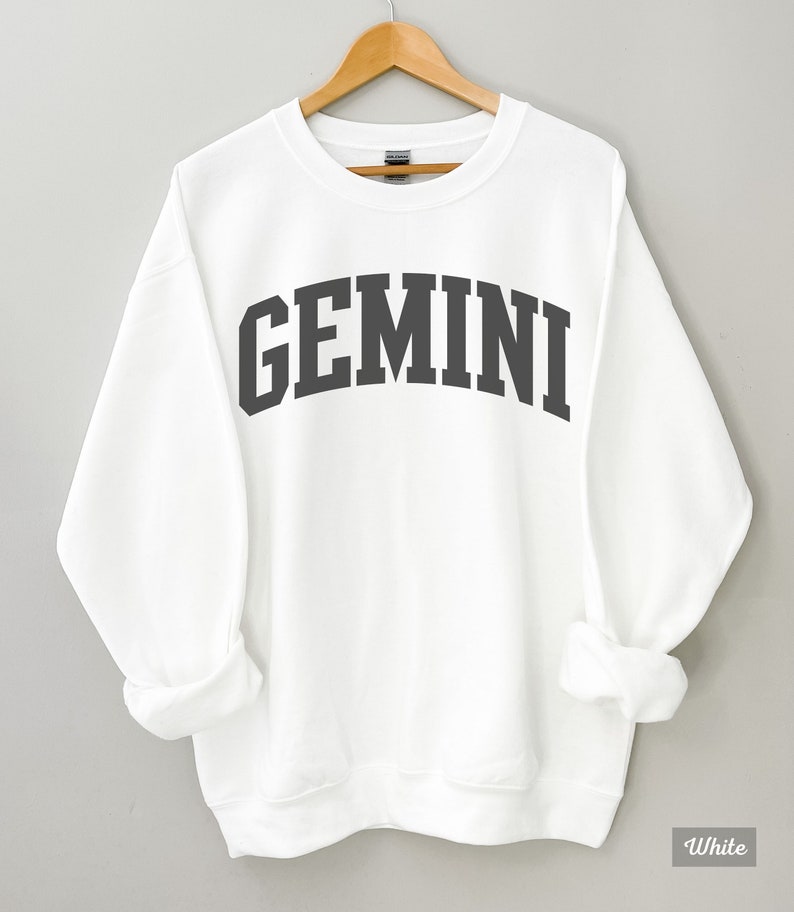 以下が含まれることがあります： 白地に黒で「GEMINI」とプリントされたスウェットシャツ。大学風のフォントを使用しています。
