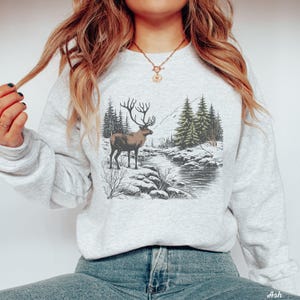May include: A gray sweatshirt with a graphic print of a deer standing in a snowy forest scene. The deer is brown with antlers and is surrounded by pine trees and a river.