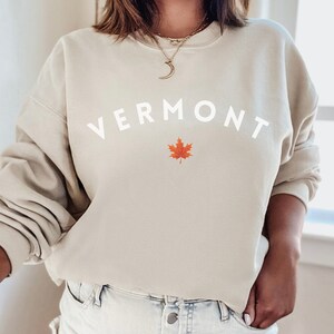 May include: A beige sweatshirt with the word "VERMONT" in white lettering and a red maple leaf graphic.