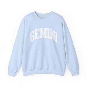 May include: A pale blue crew neck sweatshirt featuring the word "GEMINI" in white, arched lettering. The long-sleeved top is made of a soft material and is suitable for casual wear. The sweatshirt is part of the zodiac collection.