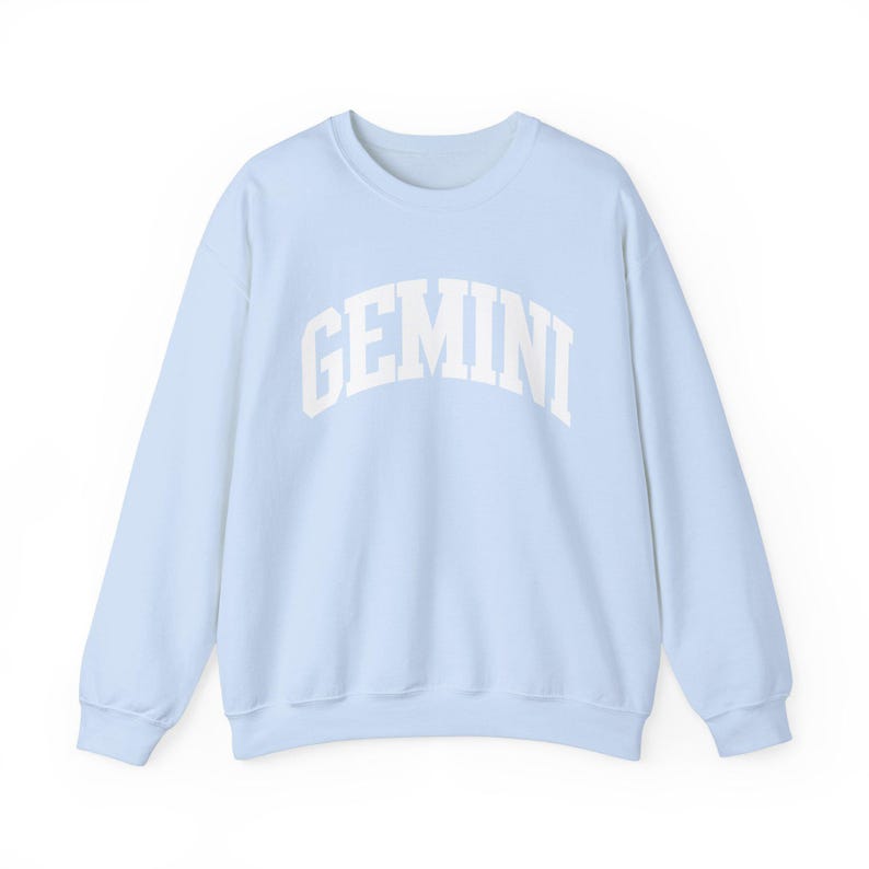 May include: A pale blue crew neck sweatshirt featuring the word "GEMINI" in white, arched lettering. The long-sleeved top is made of a soft material and is suitable for casual wear. The sweatshirt is part of the zodiac collection.