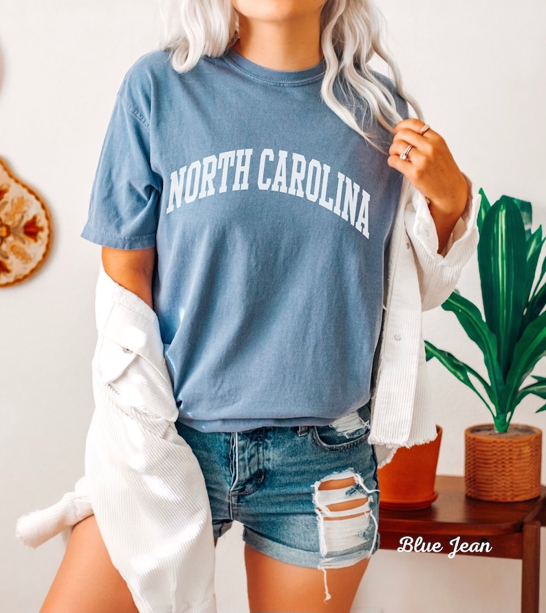 Może przedstawiać: Niebieska koszulka z napisem "NORTH CAROLINA" w kolorze białym. Koszulkę nosi osoba w białej kurtce i niebieskich dżinsowych spodenkach.