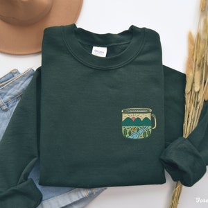 May include: Forest green sweatshirt with a pocket featuring a camping mug design with a mountain scene and a setting sun. The mug has a yellow rim and handle.
