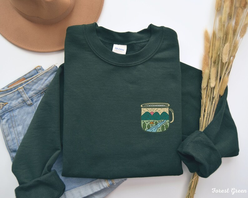 May include: Forest green sweatshirt with a pocket featuring a camping mug design with a mountain scene and a setting sun. The mug has a yellow rim and handle.
