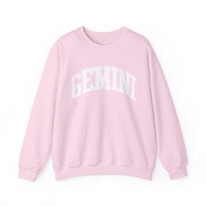 May include: A pale pink sweatshirt featuring the word "GEMINI" in white, arched lettering. This long-sleeved top is made from a soft fabric, ideal for everyday wear. The design is simple and clean.