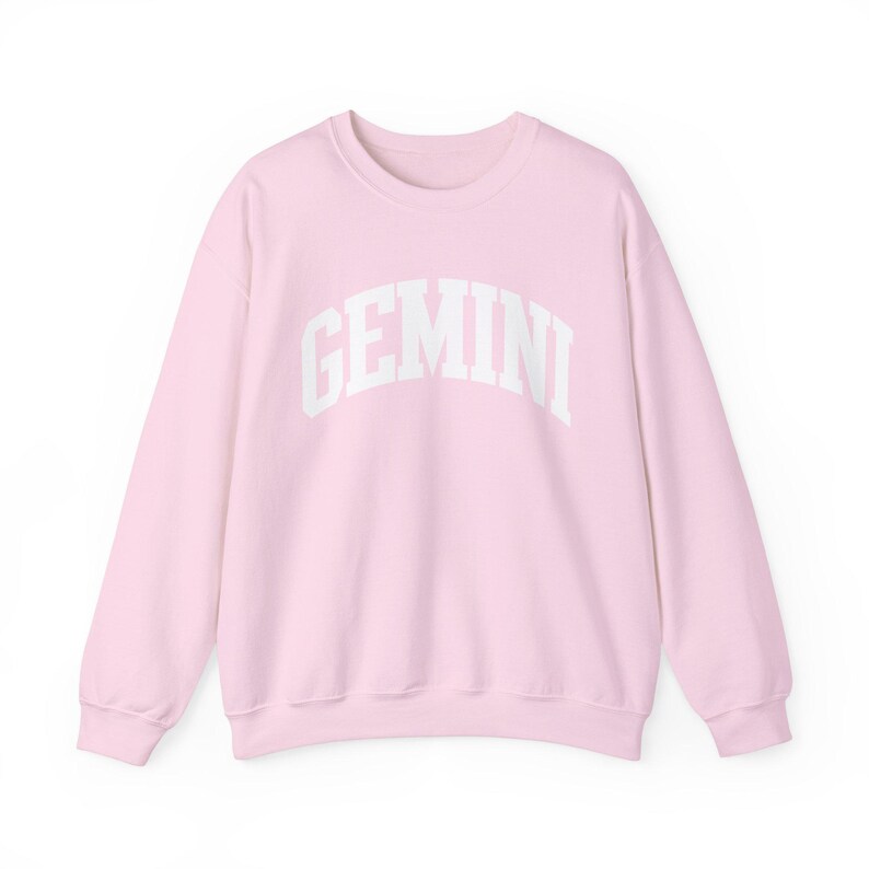 May include: A pale pink sweatshirt featuring the word "GEMINI" in white, arched lettering. This long-sleeved top is made from a soft fabric, ideal for everyday wear. The design is simple and clean.