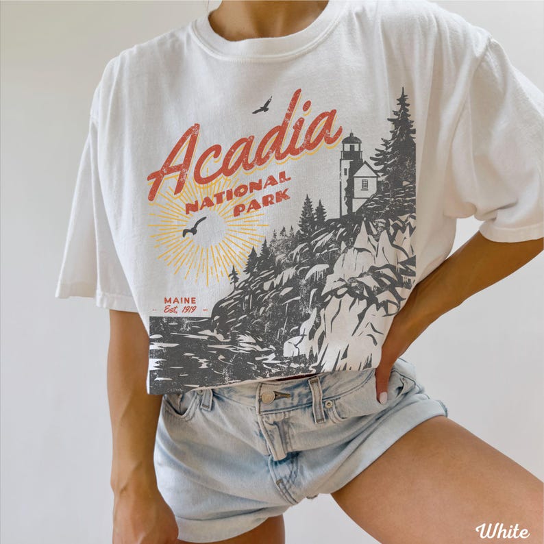 May include: White cropped t-shirt with a vintage graphic design featuring the text "Acadia National Park" and a lighthouse. The graphic also includes a sun, birds, and trees. The text "Maine Est. 1919" is also included in the design.