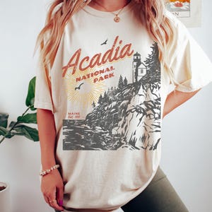May include: A cream-colored t-shirt with a vintage-style graphic of Acadia National Park in Maine. The graphic features a lighthouse, trees, and a rocky coastline. The text "Acadia National Park" is in red and yellow, and the text "Maine Est. 1919" is in black.