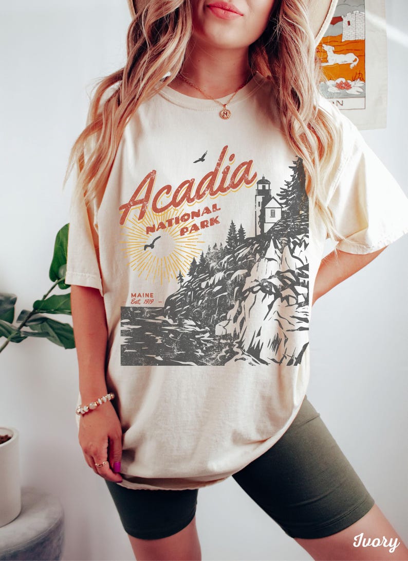 May include: A cream-colored t-shirt with a vintage-style graphic of Acadia National Park in Maine. The graphic features a lighthouse, trees, and a rocky coastline. The text "Acadia National Park" is in red and yellow, and the text "Maine Est. 1919" is in black.