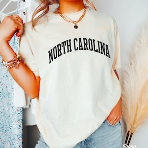 Może przedstawiać: Kremowa koszulka z napisem "NORTH CAROLINA" w kolorze czarnym, w łukowatych literach. Koszulkę nosi osoba z długimi brązowymi włosami i złotym łańcuszkiem.