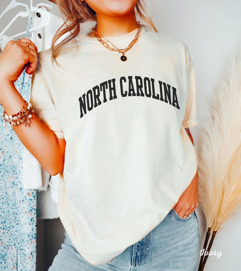 Może przedstawiać: Kremowa koszulka z napisem "NORTH CAROLINA" w kolorze czarnym, w łukowatych literach. Koszulkę nosi osoba z długimi brązowymi włosami i złotym łańcuszkiem.