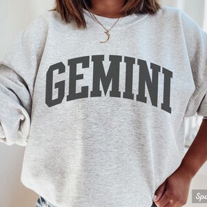 以下が含まれることがあります： グレーのトレーナーに、黒い文字で「GEMINI」とプリントされています。トレーナーはゆったりとしたフィット感で、クルーネックです。
