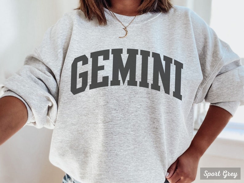 以下が含まれることがあります： グレーのトレーナーに、黒い文字で「GEMINI」とプリントされています。トレーナーはゆったりとしたフィット感で、クルーネックです。