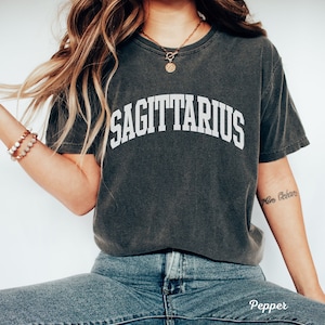 May include: A dark gray t-shirt with the word "SAGITTARIUS" printed in white on the front. The shirt is worn by a person with long blonde hair and a gold necklace. The person is wearing blue jeans.