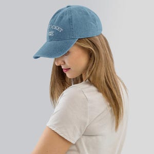 May include: A blue denim baseball cap with white embroidered text that reads "The Bucket List".