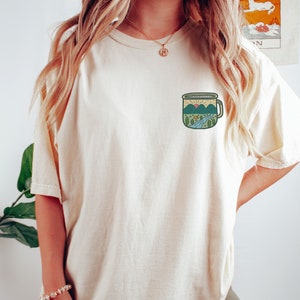 May include: A white t-shirt with a graphic of a camping mug with a mountain scene inside. The mug is green, blue, and yellow.