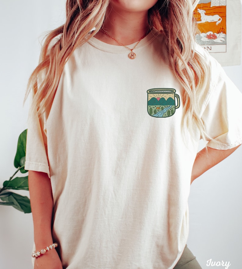 May include: A white t-shirt with a graphic of a camping mug with a mountain scene inside. The mug is green, blue, and yellow.