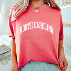 Może przedstawiać: R&oacute;żowy T-shirt z napisem "NORTH CAROLINA" w kolorze białym, w łukowatych literach.