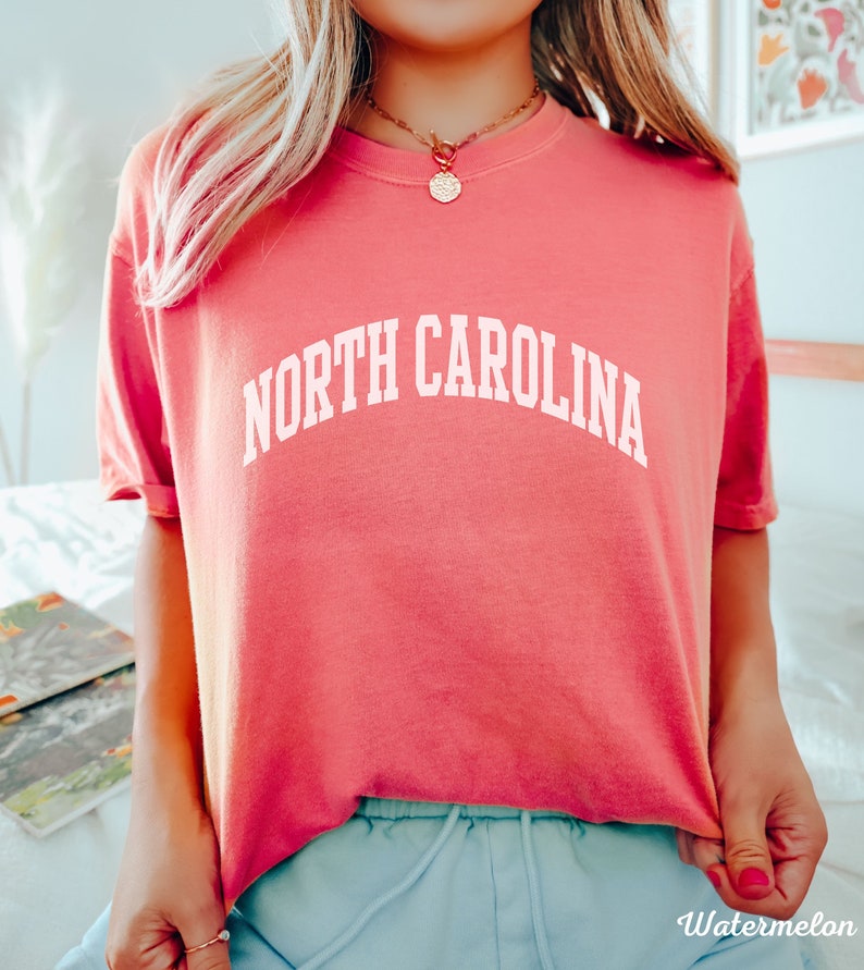 Może przedstawiać: R&oacute;żowy T-shirt z napisem "NORTH CAROLINA" w kolorze białym, w łukowatych literach.