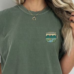 May include: A green t-shirt with a graphic of a camping mug with a mountain scene inside. The mug has a sunrise, a river, trees, and a tent.