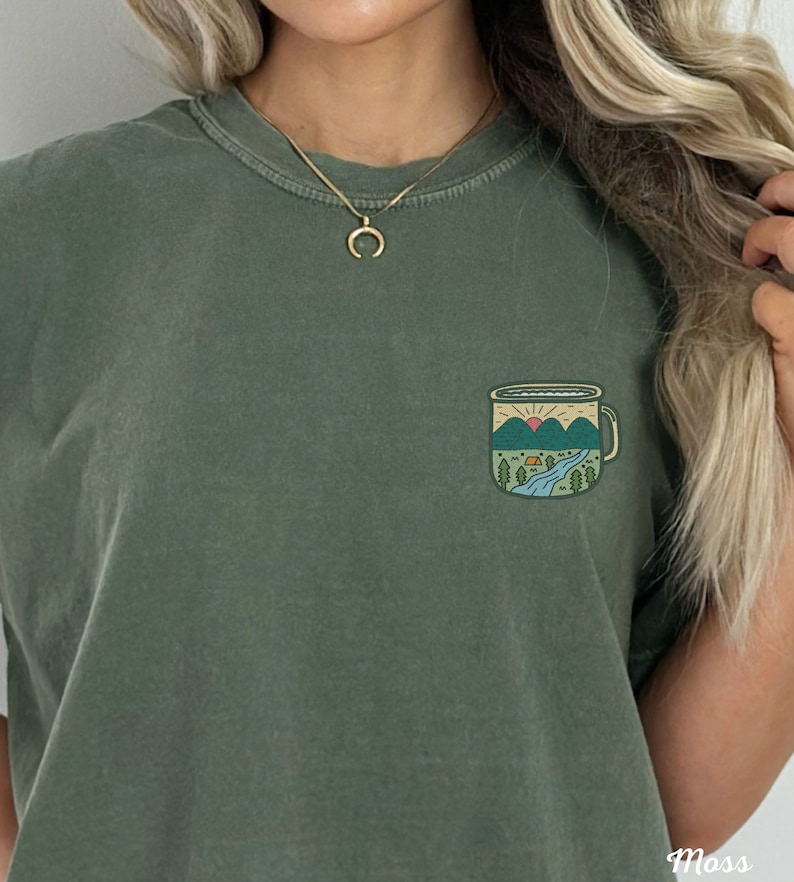 May include: A green t-shirt with a graphic of a camping mug with a mountain scene inside. The mug has a sunrise, a river, trees, and a tent.