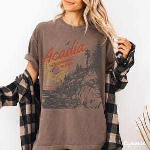 May include: A brown t-shirt with a vintage-style graphic of Acadia National Park, Maine. The graphic features a lighthouse, a rocky coastline, and the text "Acadia National Park".