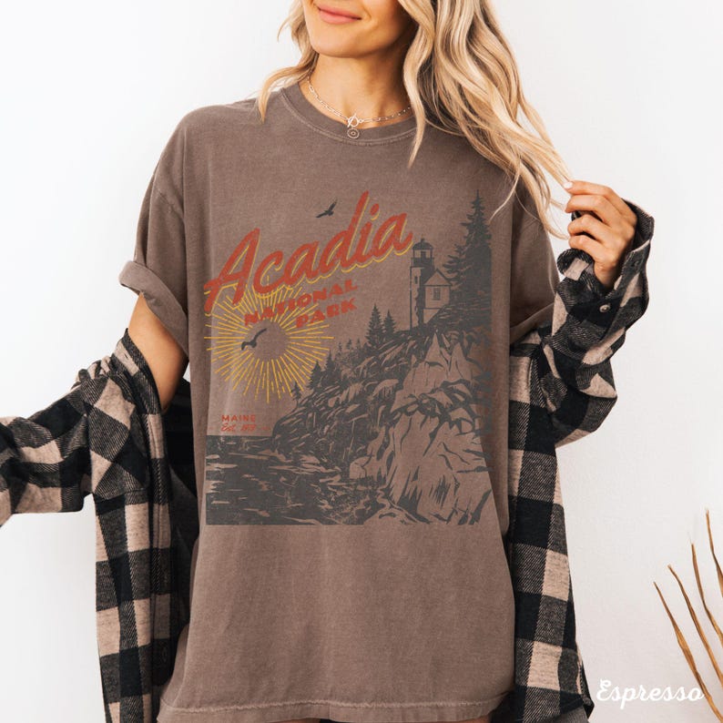 May include: A brown t-shirt with a vintage-style graphic of Acadia National Park, Maine. The graphic features a lighthouse, a rocky coastline, and the text "Acadia National Park".