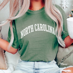 Może przedstawiać: Zielony T-shirt z napisem "NORTH CAROLINA" w kolorze białym, w łukowatych literach. Koszulkę nosi osoba z blond włosami i niebieskimi dżinsami.