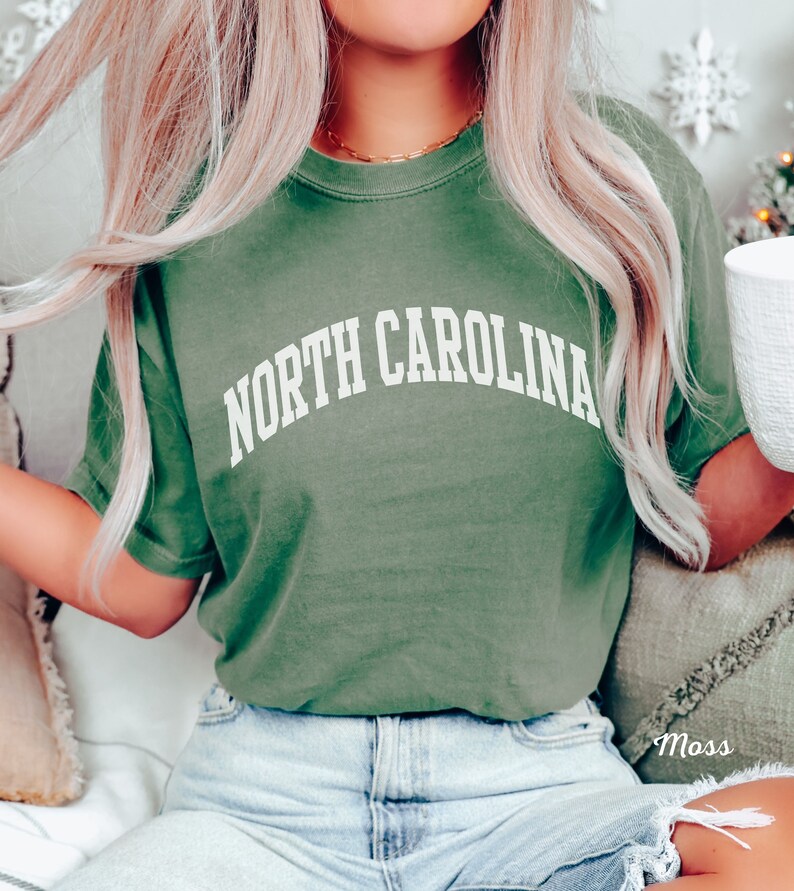 Może przedstawiać: Zielony T-shirt z napisem "NORTH CAROLINA" w kolorze białym, w łukowatych literach. Koszulkę nosi osoba z blond włosami i niebieskimi dżinsami.
