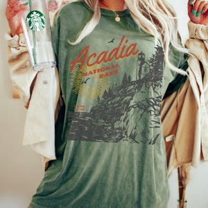 May include: Green oversized t-shirt with a vintage-style graphic of Acadia National Park, Maine. The graphic features a lighthouse, trees, and a sun with rays. The text "Acadia National Park" is in red and orange.