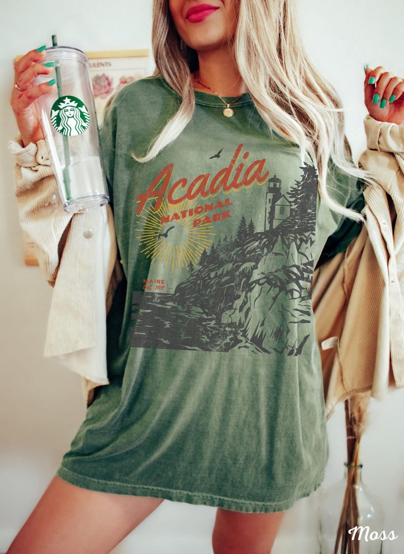 May include: Green oversized t-shirt with a vintage-style graphic of Acadia National Park, Maine. The graphic features a lighthouse, trees, and a sun with rays. The text "Acadia National Park" is in red and orange.