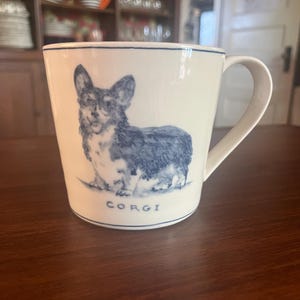 K&ouml;nnte beinhalten: Wei&szlig;e Keramiktasse mit einer blauen Illustration eines Corgi-Hundes. Die Tasse hat einen blauen Rand und einen Henkel. Das Wort "CORGI" ist blau am Boden der Tasse aufgedruckt. Die Tasse steht auf einer braunen Oberfl&auml;che.