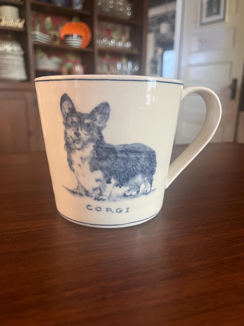 K&ouml;nnte beinhalten: Wei&szlig;e Keramiktasse mit einer blauen Illustration eines Corgi-Hundes. Die Tasse hat einen blauen Rand und einen Henkel. Das Wort "CORGI" ist blau am Boden der Tasse aufgedruckt. Die Tasse steht auf einer braunen Oberfl&auml;che.