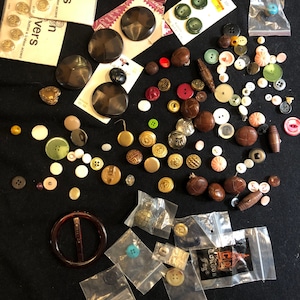May include: A collection of assorted buttons in various colors, shapes, and sizes, including brown leather buttons, gold metal buttons, and plastic buttons. Some buttons are in small plastic bags. The buttons are on a black surface.