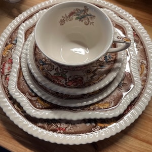 May include: A set of three white ceramic plates with a brown and floral pattern. The plates are stacked on top of each other, with a white teacup with a brown and floral pattern on top of the plates.