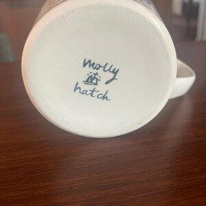 K&ouml;nnte beinhalten: Wei&szlig;er Keramikbecher mit blau-wei&szlig;em Design. Der Boden des Bechers tr&auml;gt den handgeschriebenen Text "molly hatch" in schwarzer Tinte. Der Becher hat einen Henkel und steht auf einer dunkelbraunen Holzoberfl&auml;che.