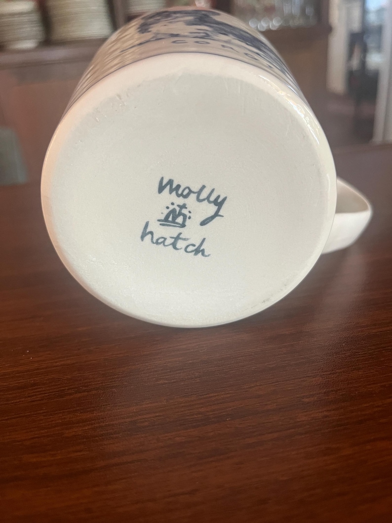 K&ouml;nnte beinhalten: Wei&szlig;er Keramikbecher mit blau-wei&szlig;em Design. Der Boden des Bechers tr&auml;gt den handgeschriebenen Text "molly hatch" in schwarzer Tinte. Der Becher hat einen Henkel und steht auf einer dunkelbraunen Holzoberfl&auml;che.
