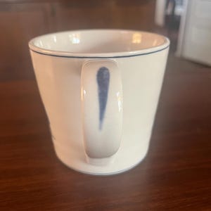 K&ouml;nnte beinhalten: Wei&szlig;e Keramiktasse mit einer blauen Linie am Rand und einem blauen Design am Griff. Die Tasse hat eine leicht gebogene Form und steht auf einer dunklen Holzoberfl&auml;che. Die Tasse ist leer.