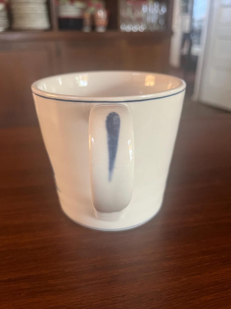 K&ouml;nnte beinhalten: Wei&szlig;e Keramiktasse mit einer blauen Linie am Rand und einem blauen Design am Griff. Die Tasse hat eine leicht gebogene Form und steht auf einer dunklen Holzoberfl&auml;che. Die Tasse ist leer.