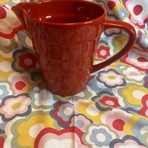 May include: A red ceramic pitcher with a textured design. The pitcher has a handle and a spout.