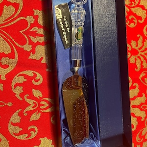 May include: A Waterford Crystal cake server in a blue presentation box. The server has a clear crystal handle with a silver-tone metal base and a gold-colored blade. A tag reads "Caring for Waterford Crystal."