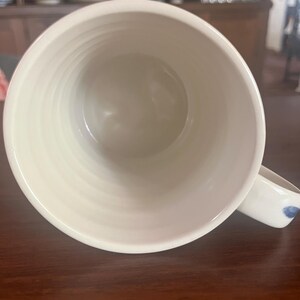 K&ouml;nnte beinhalten: Cremefarbene Keramiktasse mit breiter &Ouml;ffnung und Henkel. Der Innenraum ist glatt, hellbeige, w&auml;hrend die Au&szlig;enseite ein strukturiertes, geriffeltes Design aufweist. Der Henkel hat ein blaues Blumenmuster. Ideal f&uuml;r Tee oder Kaffee.