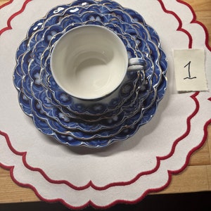May include: A set of blue and gold porcelain teacups and saucers with a floral pattern. The teacups and saucers are on a white doily with a red scalloped edge.
