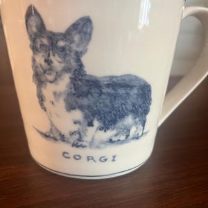 K&ouml;nnte beinhalten: Wei&szlig;e Keramik-Tasse mit einer blauen Illustration eines Corgi-Hundes. Die Tasse hat einen Henkel und einen blauen Rand am oberen Rand. Das Wort "CORGI" ist blau unter dem Hund aufgedruckt.
