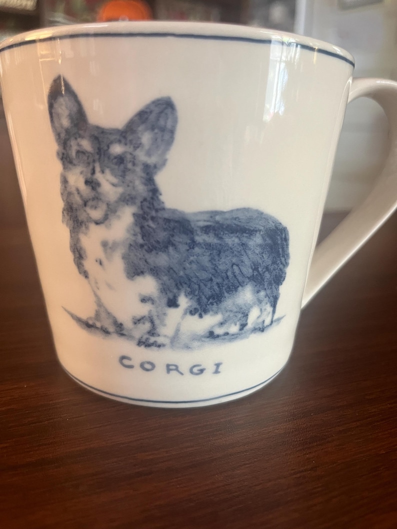 K&ouml;nnte beinhalten: Wei&szlig;e Keramik-Tasse mit einer blauen Illustration eines Corgi-Hundes. Die Tasse hat einen Henkel und einen blauen Rand am oberen Rand. Das Wort "CORGI" ist blau unter dem Hund aufgedruckt.