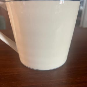 K&ouml;nnte beinhalten: Wei&szlig;e Keramiktasse mit schlichtem Design. Die Tasse hat einen Henkel und einen d&uuml;nnen blauen Strich um den Rand und die Basis. Die Tasse steht auf einer dunkelbraunen Holzoberfl&auml;che.