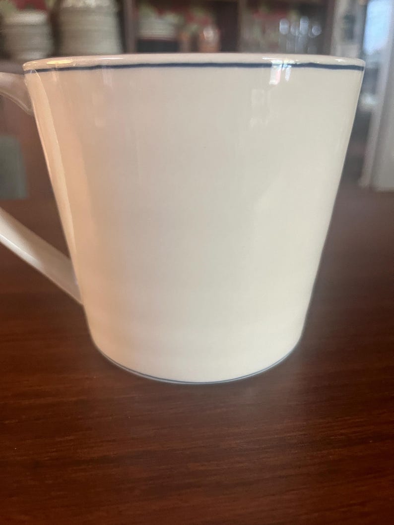 K&ouml;nnte beinhalten: Wei&szlig;e Keramiktasse mit schlichtem Design. Die Tasse hat einen Henkel und einen d&uuml;nnen blauen Strich um den Rand und die Basis. Die Tasse steht auf einer dunkelbraunen Holzoberfl&auml;che.