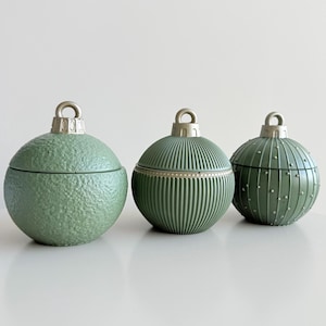 May include: Three green, ball-shaped decorative containers with silver metal loops. The containers have varied textures: one is textured, one has vertical lines, and the third has small bumps. These containers are likely for decorative purposes.