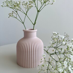 May include: A pale pink ceramic vase with vertical ridges, holding a bouquet of white baby's breath flowers.